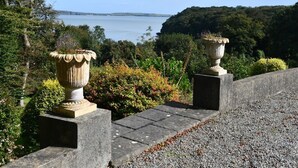 Un chemin de pierre bordé de deux grands vases, entouré d’une végétation luxuriante et offrant une vue sur une étendue d’eau.