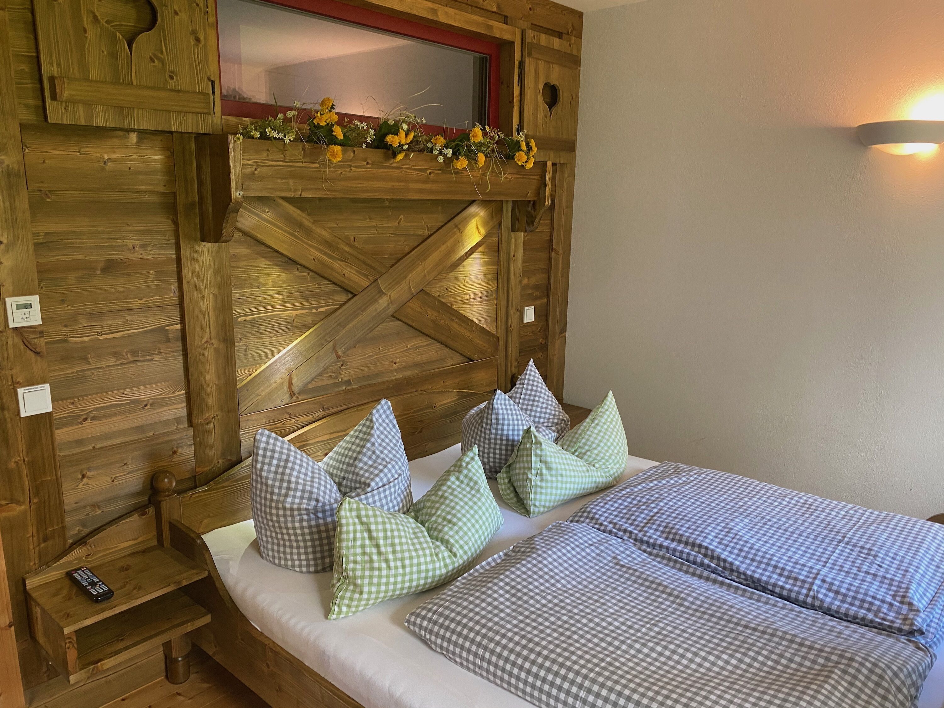 Un lit en bois avec une literie à carreaux et des oreillers, une petite table de chevet avec une télécommande et un luminaire fixé au mur.