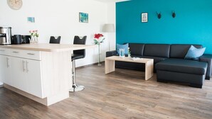 A modern living room with a black sofa, a coffee table, and a kitchen area with a white cabinet and a clock. - Ferienhof - Weites - Land, large apartment with terrace, garden and children's playground (Stadtkyll)