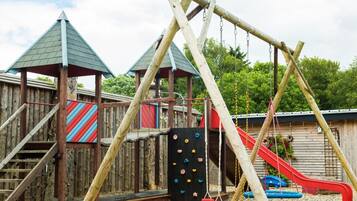 Ein Spielplatz mit Holzkonstruktionen, darunter eine Rutsche und eine Kletterwand, umzäunt durch einen Holzzaun.