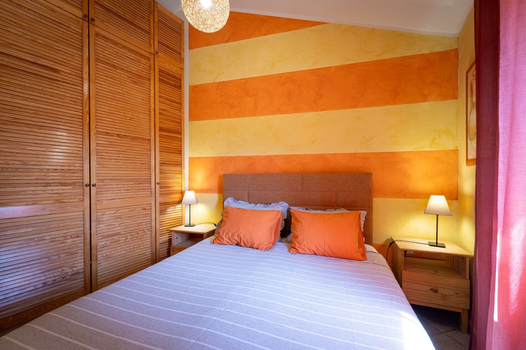 A bedroom with a striped bedspread, two nightstands with lamps, a wooden wardrobe, and a wall with horizontal stripes.