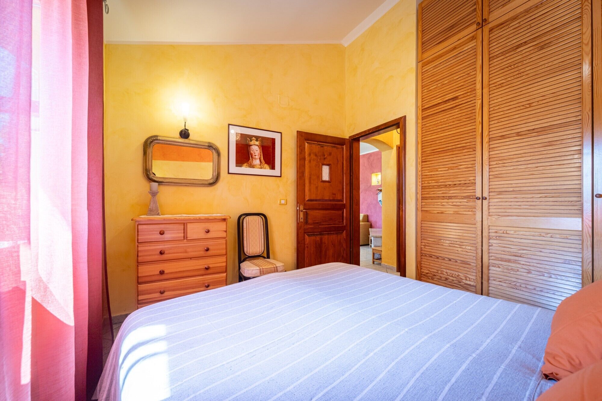 A bedroom with a bed, wooden wardrobe, a dresser, a mirror, and a framed picture on the wall.