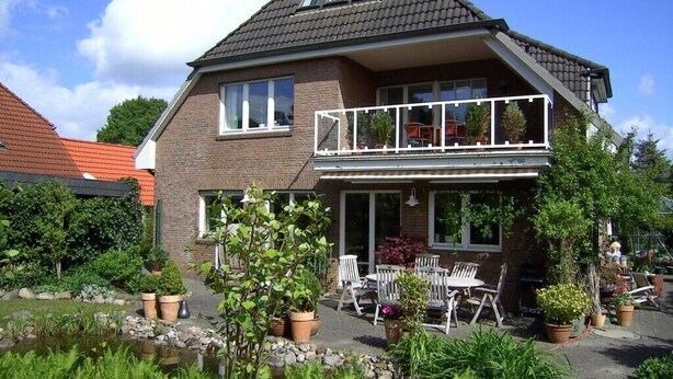 Uma casa de dois andares com varanda, jardim com diversas plantas e móveis de exterior.