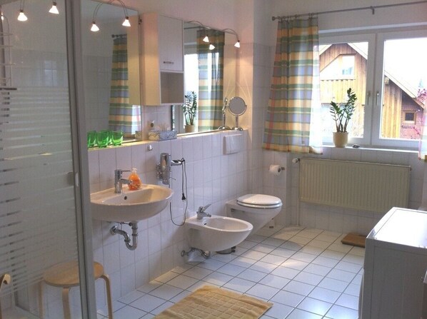A bathroom with a sink, bidet, toilet, and a mirror. There is a window with curtains and a radiator. - beautiful, high-quality holiday flat, 3 rooms, near the Kieler fjord (Flintbek)