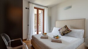 Un lit bien fait, avec du linge de lit blanc, une chaise en plastique transparent et un mur de pierre visible à travers la fenêtre.