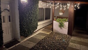 Un sendero conduce a un espacio al aire libre techado, con luces colgantes, una puerta blanca y una pared cubierta de follaje.