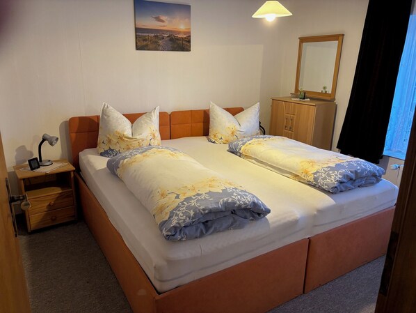 Un lit double avec une literie à motifs floraux, une table de chevet avec une lampe, une commode en bois et un tableau représentant un coucher de soleil accroché au mur.