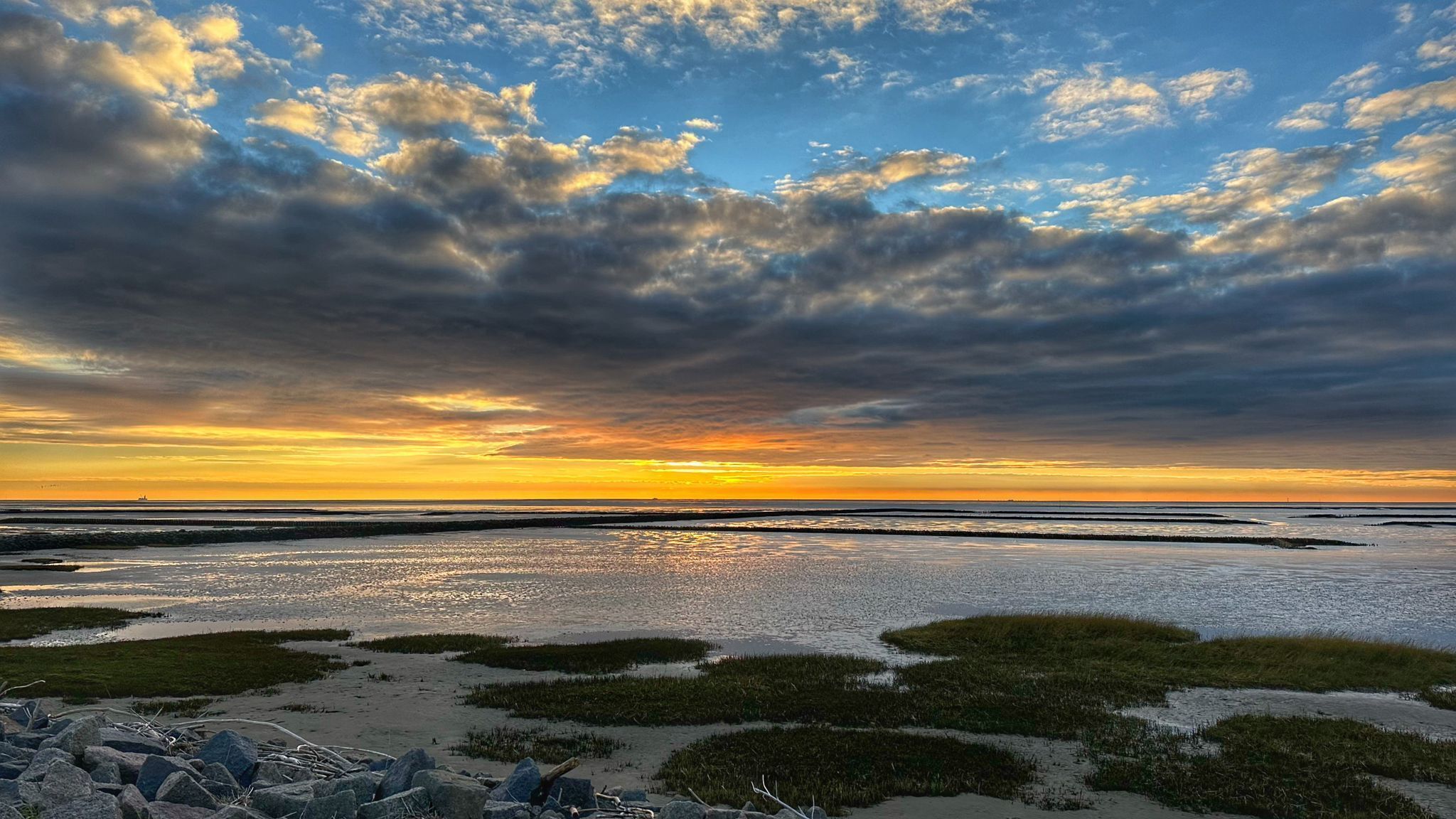 A coastal landscape with a sunset, featuring a sandy beach, patches of vegetation, and a cloudy sky.