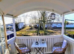 Ein überdachter Außenbereich mit zwei Korbstühlen und einem Tisch, der auf einen Hof mit kahlen Bäumen und geparkten Fahrzeugen blickt.