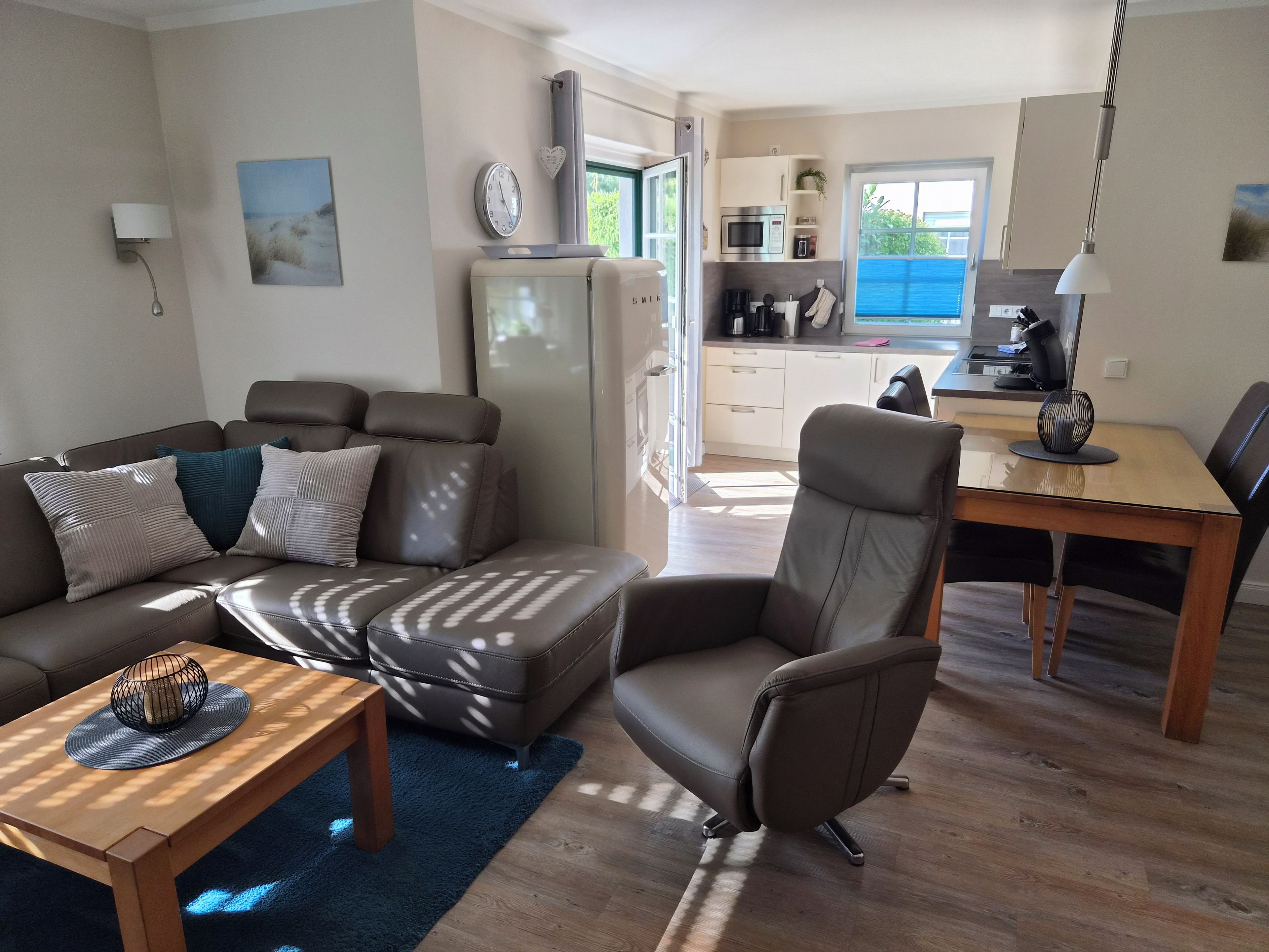 A modern living room with a sofa, armchair, and coffee table. A kitchen with appliances and a dining area is visible in the background.