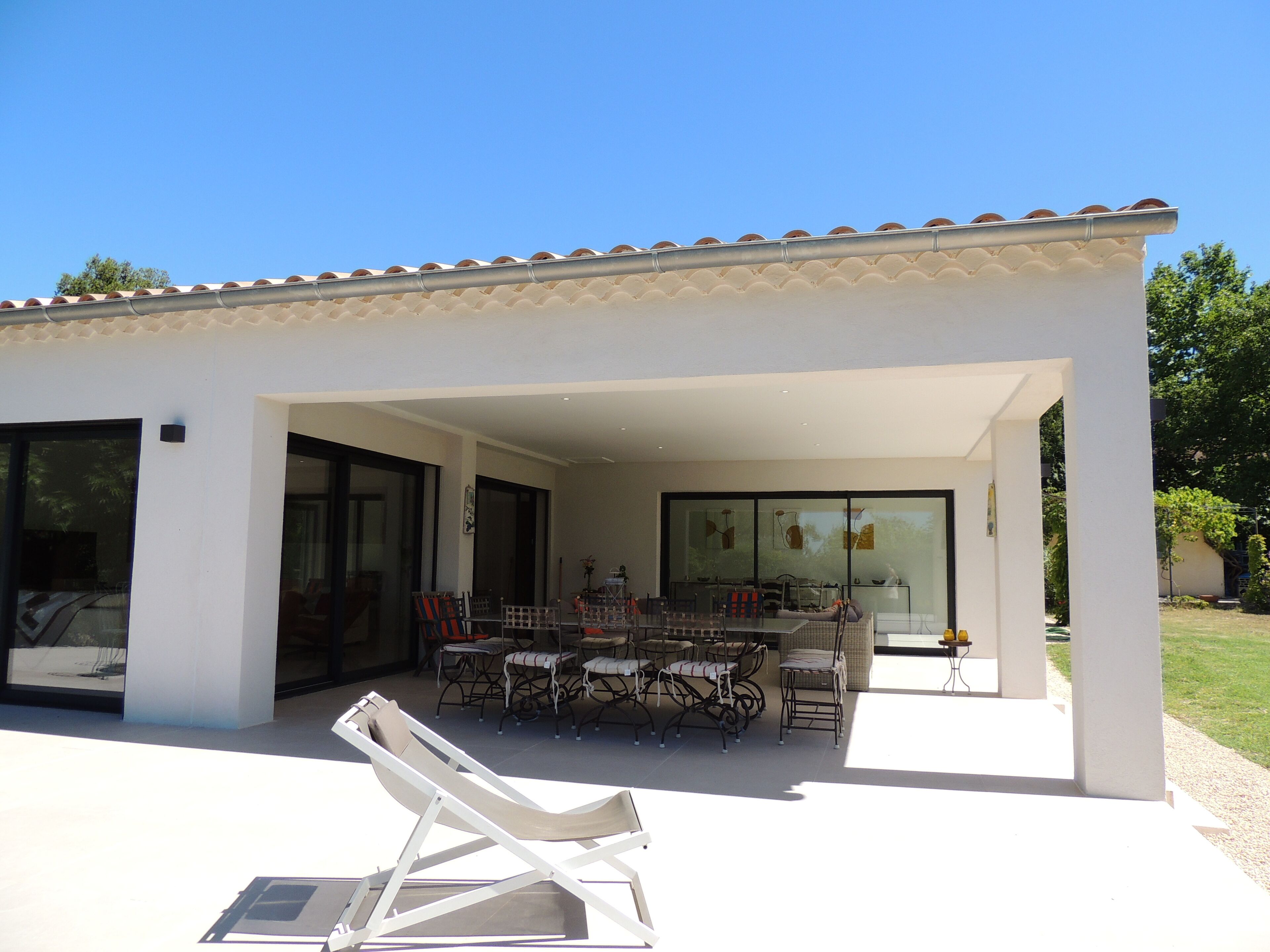 Una casa moderna con un porche cubierto, ventanas amplias y un comedor con mobiliario de exterior.