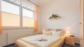 Een netjes opgemaakt bed met een opgevouwen deken en een tijdschrift, een nachtkastje met een lamp, een plank met planten, en een raam met jaloezieën.