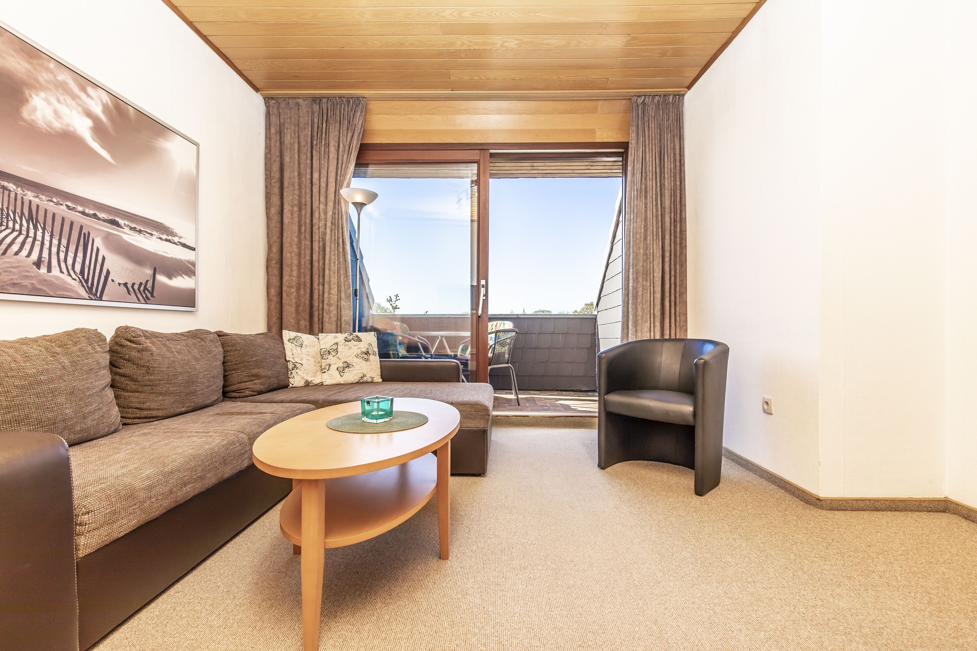 A modern living room with a sofa, a round coffee table, and a leather armchair. A large window with curtains provides a view of the outdoors.