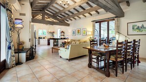A spacious living area with a wooden dining table and chairs, a sofa, and a kitchen in the background. - Private sunny villa with large heated pool, air-conditioning and river access. (Céret)