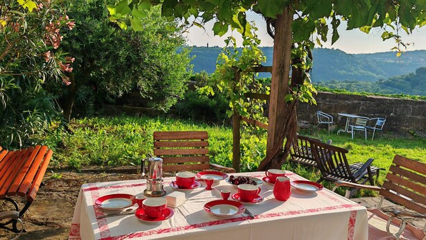 Tavolo apparecchiato all'aperto con stoviglie rosse e bianche, circondato da sedie e panche in legno, con vista su un giardino rigoglioso e dolci colline.