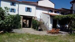 Un edificio di due piani con facciata bianca e persiane blu, un ingresso in pietra e un piccolo patio con due sedie di legno.