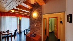 Una habitación con techo de madera, pared de acento de piedra, una zona de comedor con mesa y sillas, y un baño visible a través de una puerta abierta.