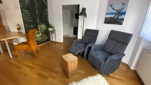 A living room with a wooden floor, two grey armchairs, a small wooden stool, a white furry rug, a dining table with a chair, a lamp, and a painting of a deer.