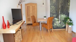 A dining area with a wooden table, two chairs, a television on a wooden stand, and a large window with a forest view.