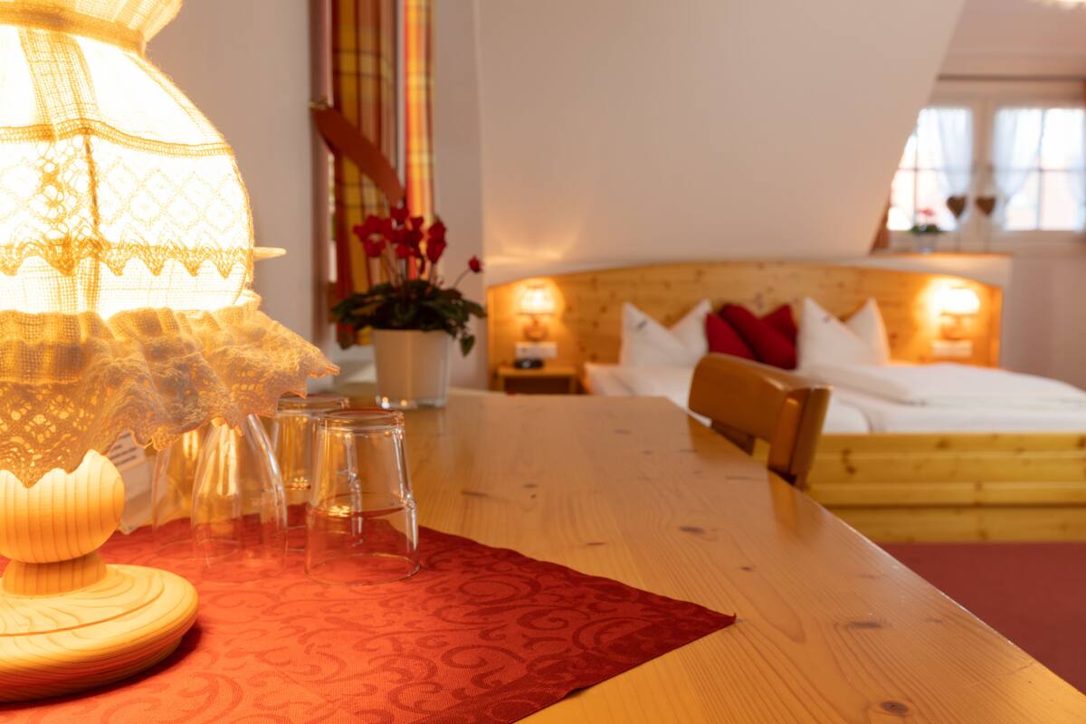 Ein Hotelzimmer mit einem hölzernen Bett, einer rot gemusterten Tischdecke, einer Lampe und einer Vase mit Blumen.