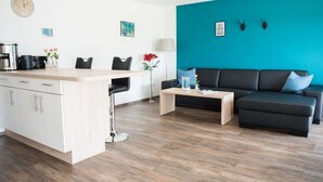A modern living room with a black sofa, a coffee table, and a kitchen area with a white cabinet and a clock. - Ferienhof - Weites - Land, large apartment with terrace, garden and children's playground (Stadtkyll)