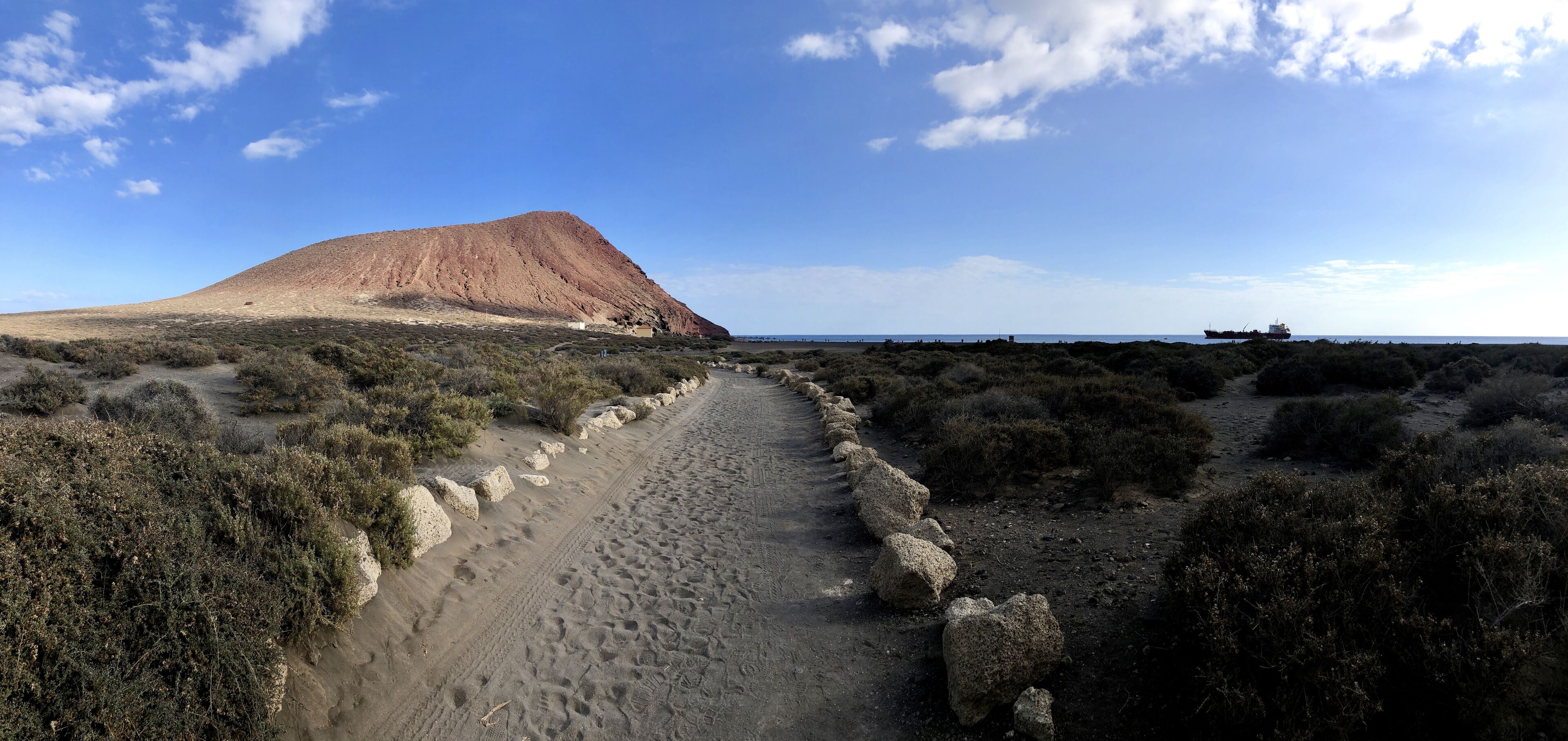 Paisaje desértico con una colina destacada, un sendero de tierra, vegetación escasa y un barco a lo lejos.