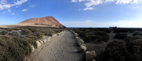 A desert landscape with a prominent hill, a dirt path, sparse vegetation, and a distant ship on the horizon.