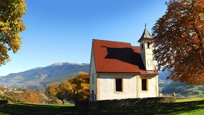 Un pequeño edificio blanco con techo rojo y un campanario, rodeado de árboles otoñales y un paisaje montañoso.