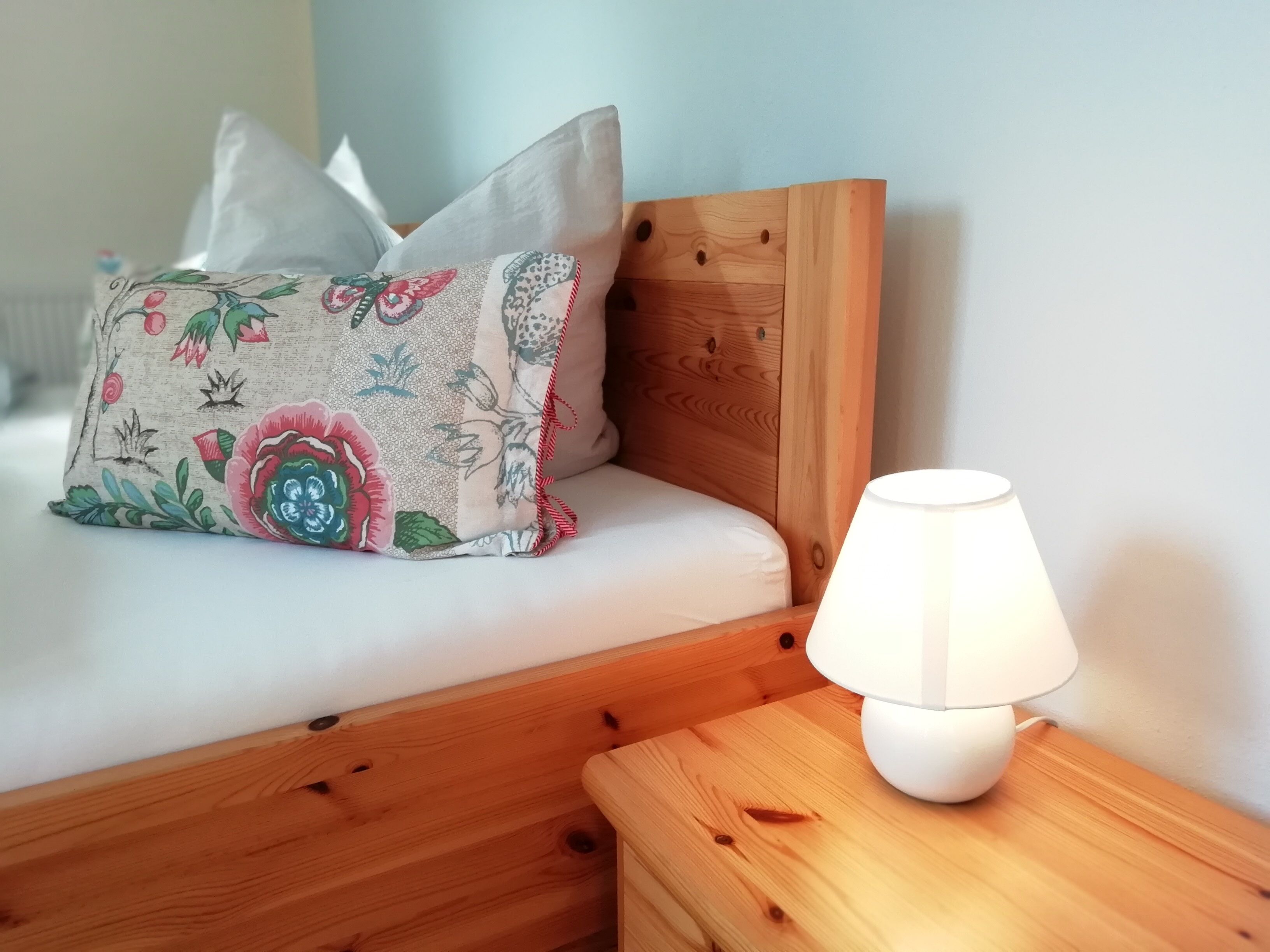A wooden bed with a floral pillow and a bedside table with a white lamp.