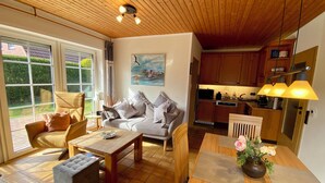 A cozy living room with a sofa, coffee table, and ottoman, a dining area with a table and chairs, and a kitchen with wooden cabinets and a sink. -  single-storey quiet apartment with garden in a central location of Greetsiel (Greetsiel)