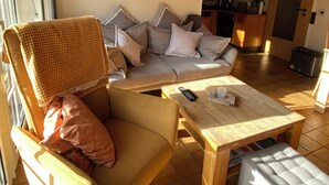 A living room with a sofa, armchair, and coffee table. There is a painting on the wall and a kitchen in the background. -  single-storey quiet apartment with garden in a central location of Greetsiel (Greetsiel)