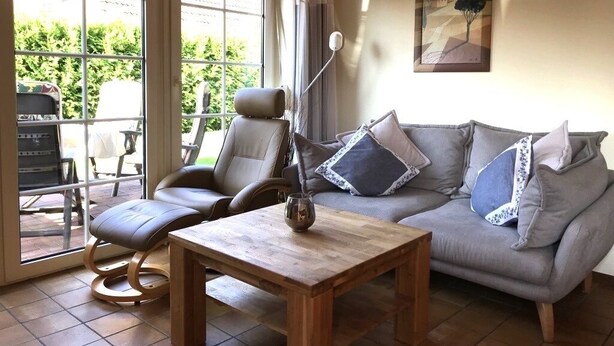 A living room with a sofa, a coffee table, and a chair. There is a window with curtains, a painting on the wall, and a view of greenery outside. -  single-storey quiet apartment with garden in a central location of Greetsiel (Greetsiel)