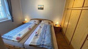 A bedroom with a wooden bed, two bedside tables with lamps, a window with curtains, a wooden wardrobe, and a small painting on the wall. -  single-storey quiet apartment with garden in a central location of Greetsiel (Greetsiel)