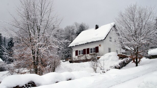Een huis bedekt met sneeuw, met een rood dak en witte muren, omringd door bomen en een besneeuwd landschap.