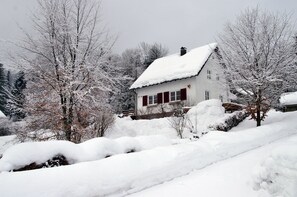 Ett snötäckt hus med ett rött tak och vita väggar, omgivet av träd och ett snöigt landskap.