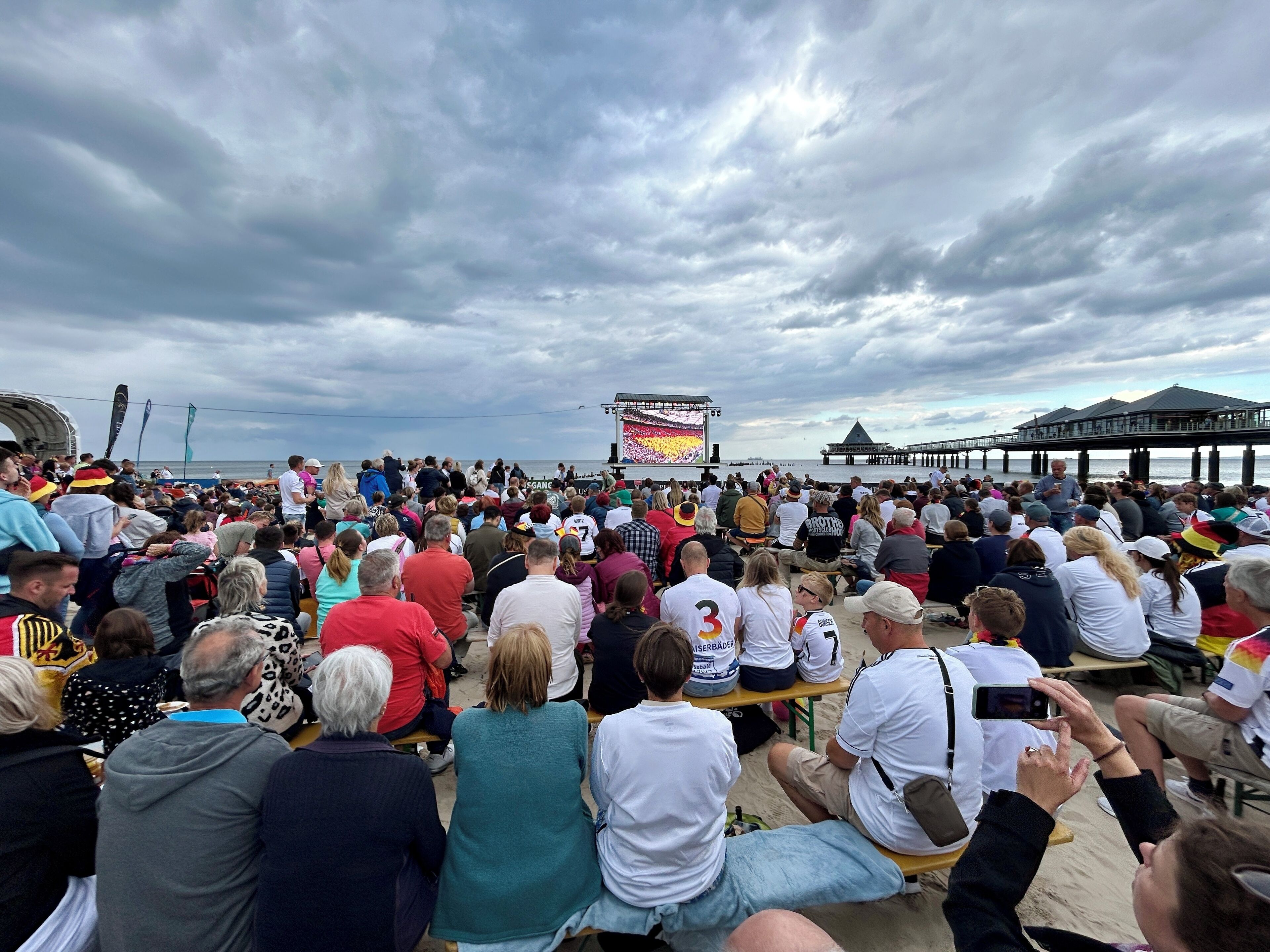 Ein belebtes Freiluft-Event mit Bühne und einer großen Leinwand, vor dem Hintergrund eines bewölkten Himmels.