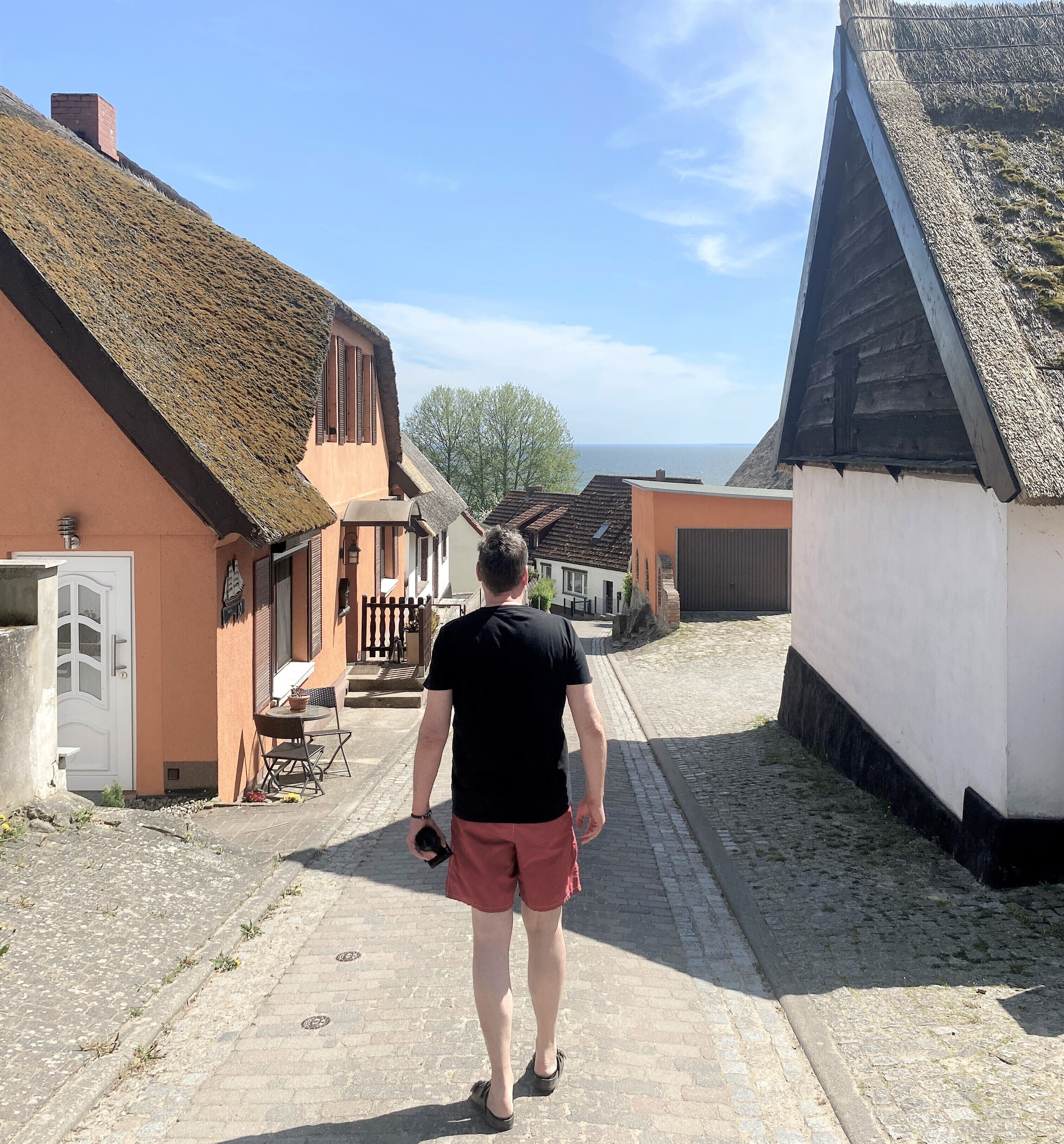 A narrow cobblestone street lined with traditional buildings, a person walking away from the camera, and a clear view of the sea in the distance.