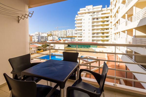 A balcony with a table and chairs, overlooking a swimming pool and adjacent buildings.