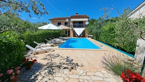 A pool area with lounge chairs and umbrellas, surrounded by greenery and a building with a tiled roof.
