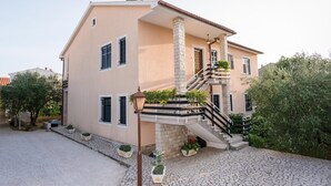 A two-story house with a stone staircase, a balcony with plants, and a paved driveway.