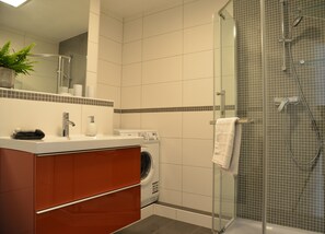 Moderni kylpyhuone, jossa on lasiseinällä varustettu suihku, sisäänrakennettu pesukone ja peilillä varustettu allaskaappi.