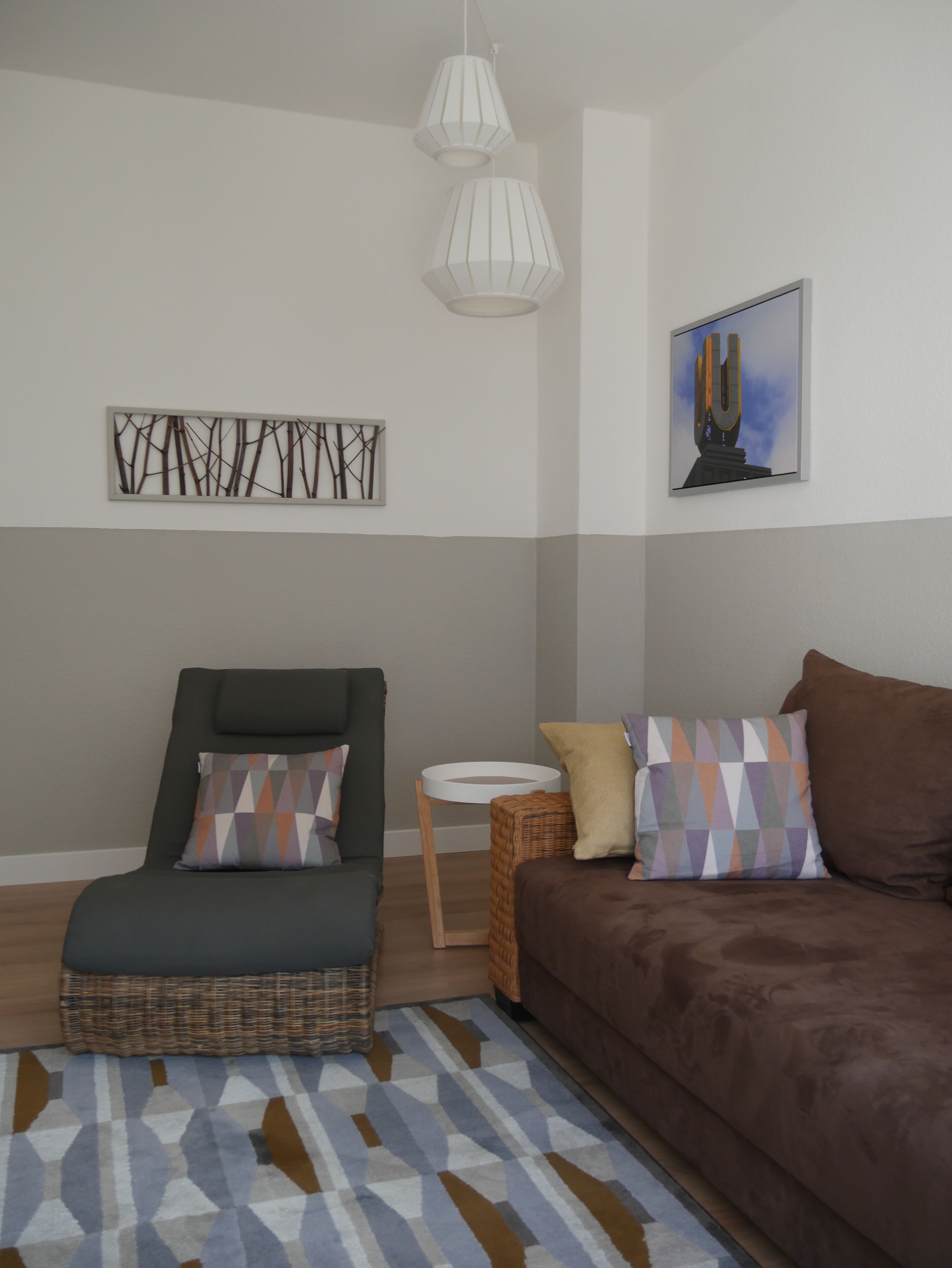 Ein modernes Wohnzimmer mit einem dunkelgrünen Sessel, einem braunen Sofa, einem gemusterten Teppich, einem kleinen runden Tisch und einem abstraktem Kunstwerk an der Wand.