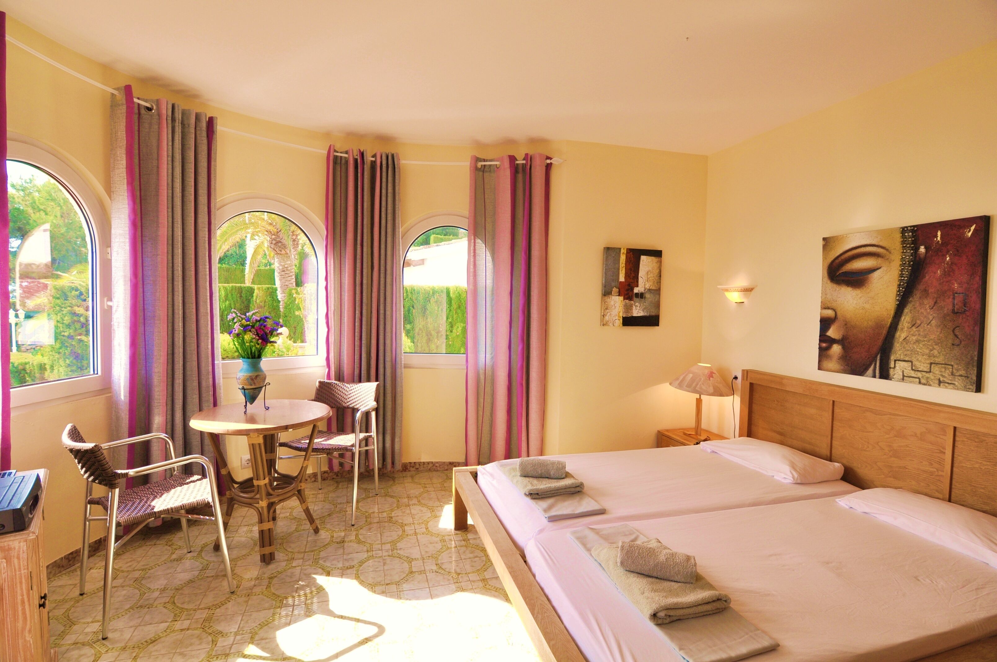 A hotel room with two beds, a dining table with chairs, a small table with a vase of flowers, and large windows with curtains.
