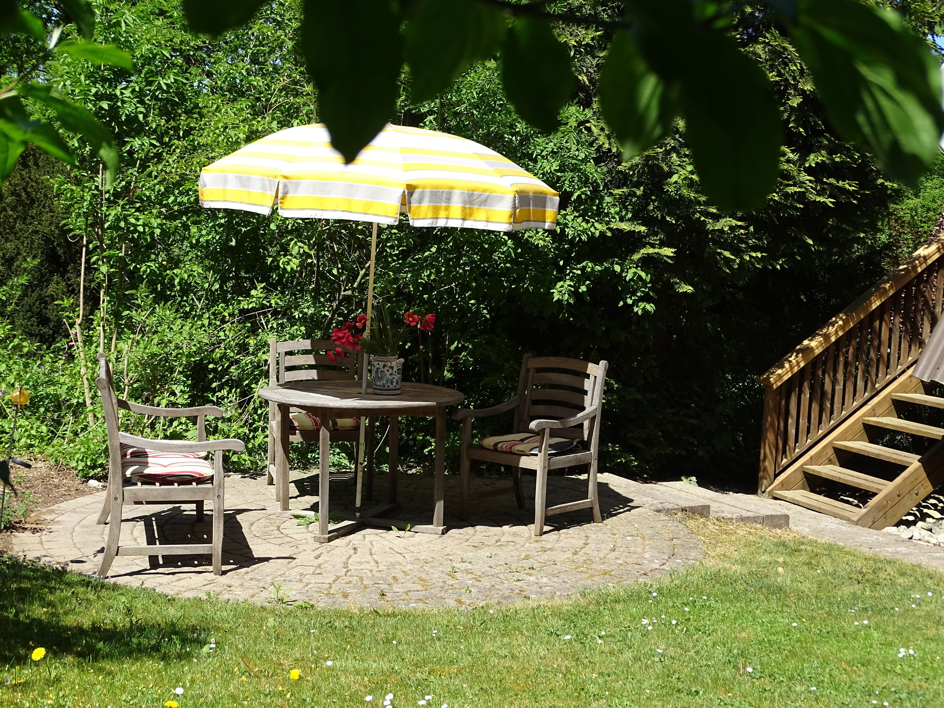 A patio with a round table and two chairs, a yellow-striped umbrella, and a potted plant with red flowers.