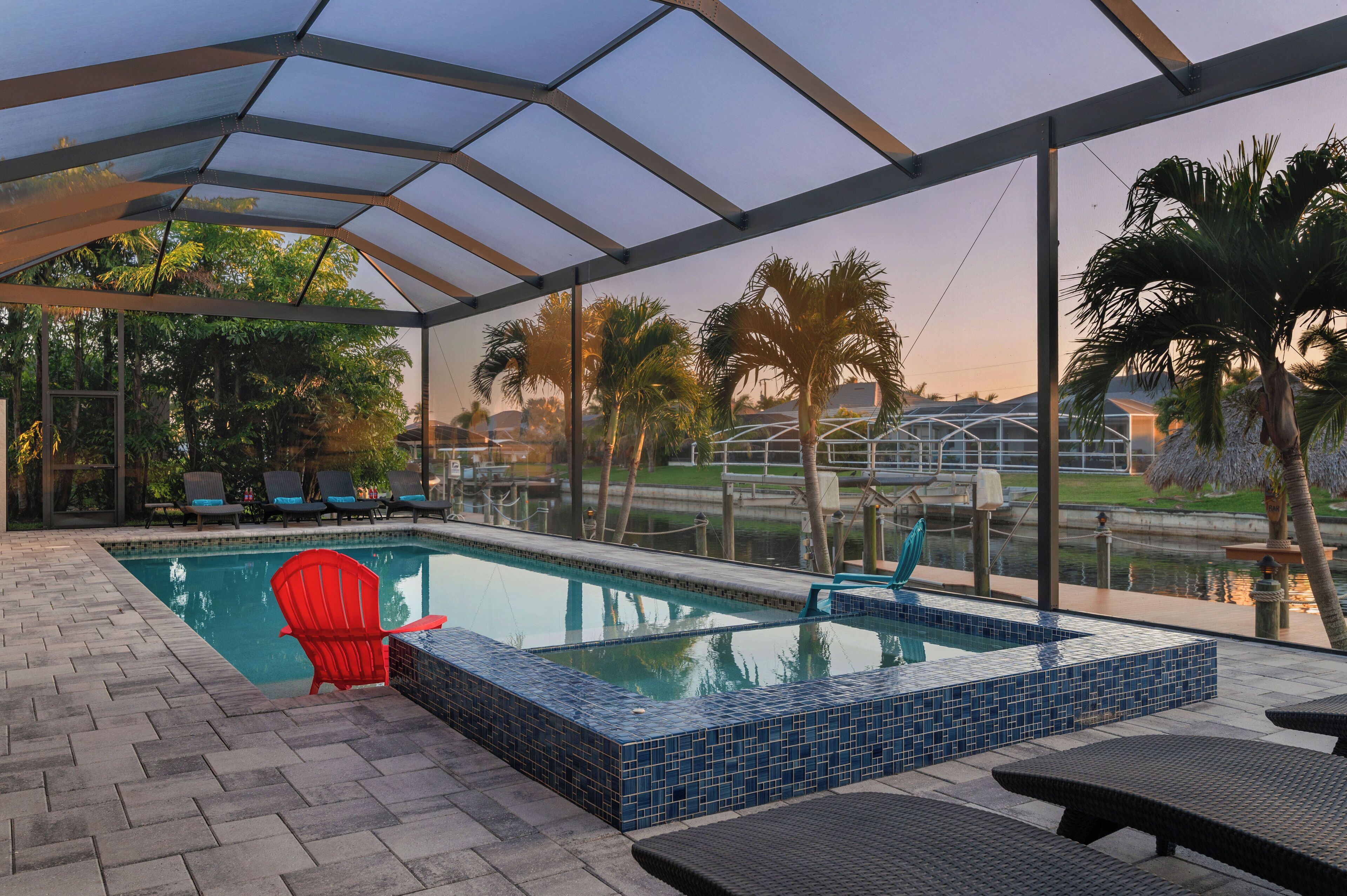 A covered outdoor swimming pool with lounge chairs and a red chair, surrounded by palm trees and a view of a canal.