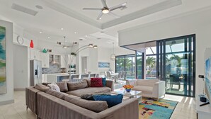 Uma sala de estar moderna com um amplo sofá modular, uma mesa de centro e um tapete colorido. Há uma área de refeições com uma mesa branca e cadeiras, e uma cozinha com armários brancos e um vaso vermelho.