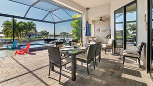 Uma área de refeições ao ar livre coberta, com vista para uma piscina e palmeiras.