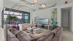 Uma sala de estar moderna com um sofá espaçoso, uma mesa de centro, uma TV e uma estante de livros. Há uma grande janela com vista para o exterior.