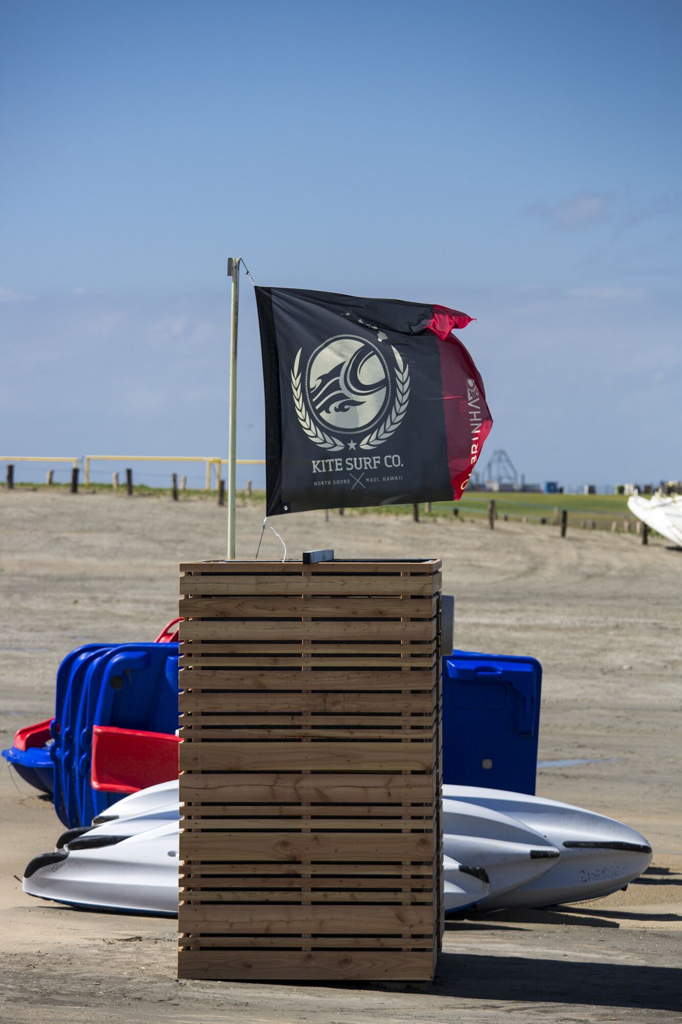 En hög med träkistor på en sandstrand, med en surfbräda och en flagga i bakgrunden.