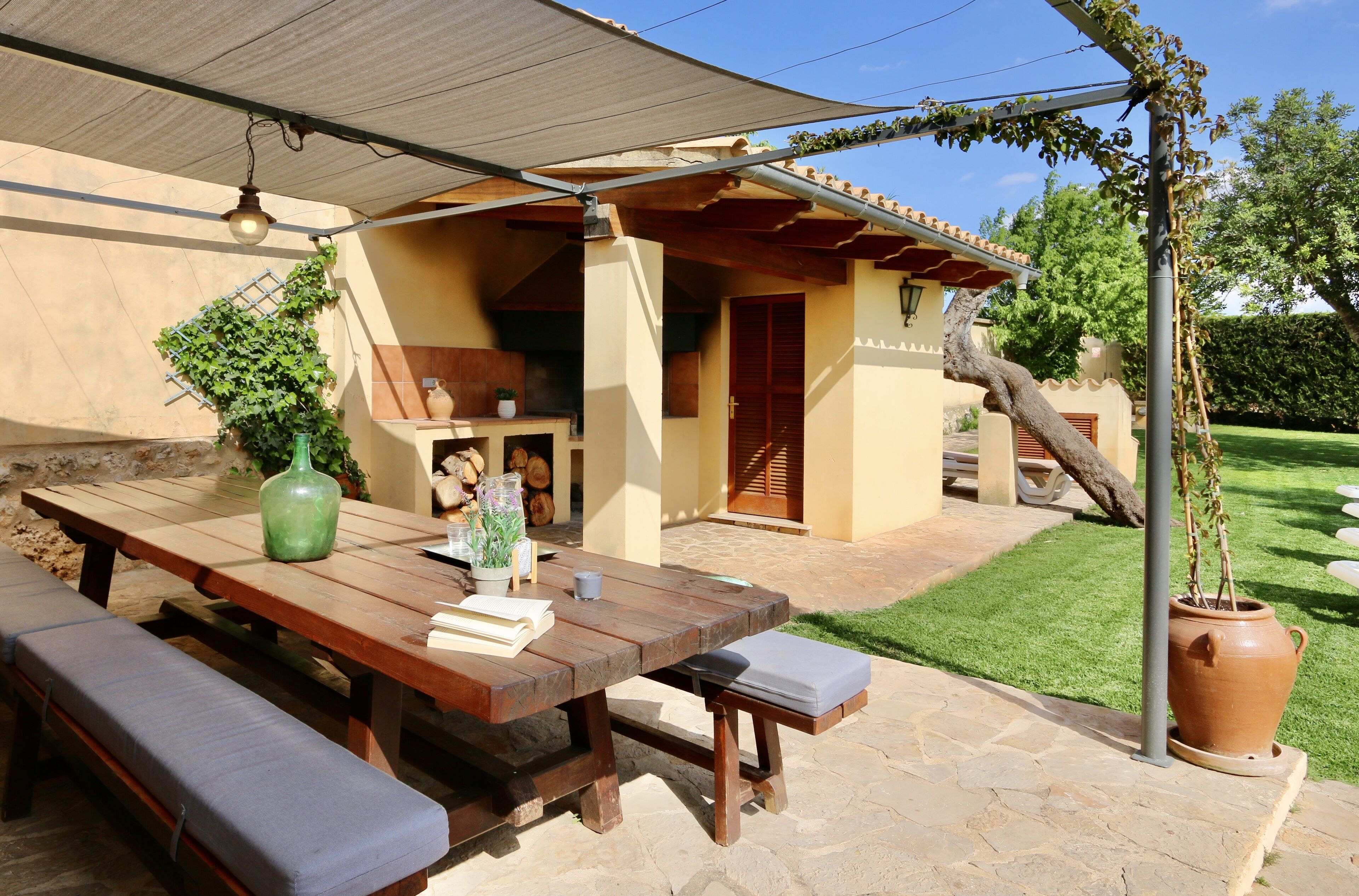 Een terras buiten met een houten tafel en banken, een pergola en een grote terracotta pot.
